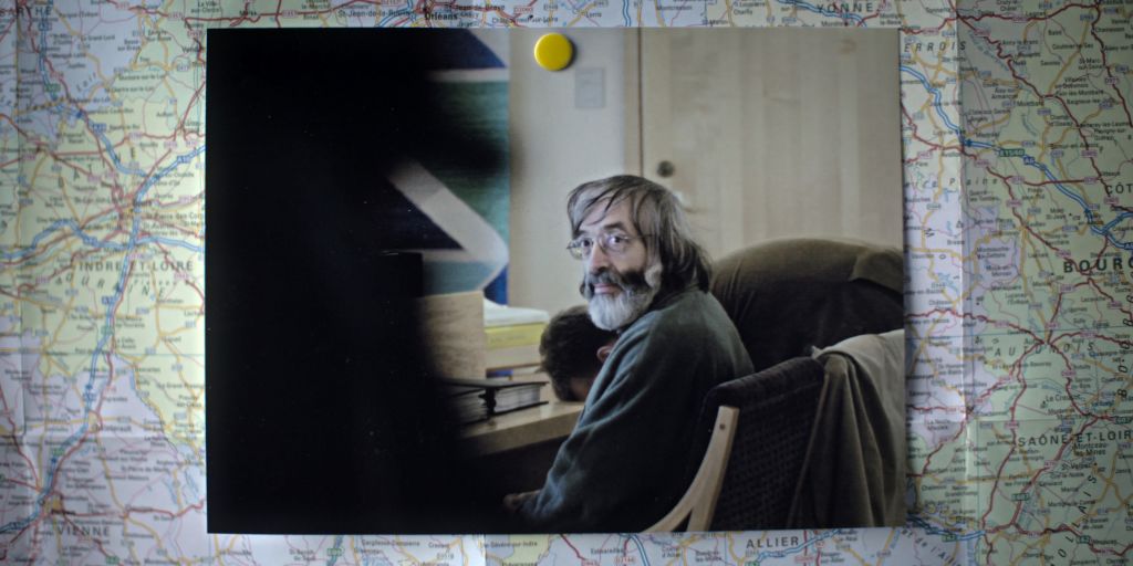 A photograph of a man is pinned atop a map. The man in the photograph is sitting in a drab courtroom and turns to face the camera. He is in his 60s and has long salt-and-pepper hair and a bushy beard. He is wearing glasses.