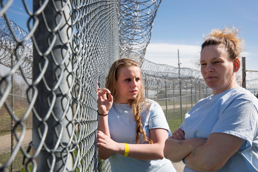 Two adult women, both white with blonde hair, stand in a prison hard by a fence. They are wearing white t-shirts.