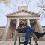 A low angle shot observes filmmaker Ric Esther Bienstock as she sits cross-legged on a chair while reading from her phone. She is a white woman white brown puffy hair, and is wearing blue jeans, a black leather jacket, and suede Chelsea boots.