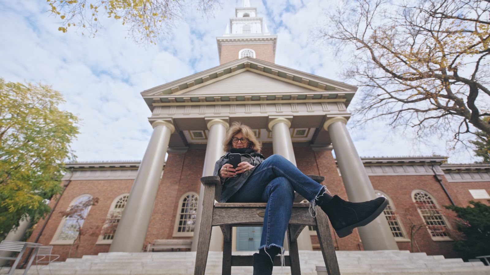 A low angle shot observes filmmaker Ric Esther Bienstock as she sits cross-legged on a chair while reading from her phone. She is a white woman white brown puffy hair, and is wearing blue jeans, a black leather jacket, and suede Chelsea boots.