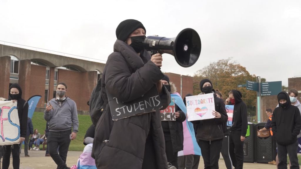 A student wearing a black puffy winter jacket, black toque, and facemask holds a megaphone during a protest.