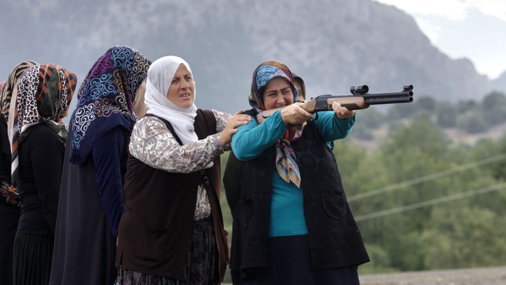 Four women practice shooting with a rifle. They are all wearing veils. One is holding the gun and pointing at a target offscreen, while a woman behind her steadies her arm.