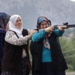Four women practice shooting with a rifle. They are all wearing veils. One is holding the gun and pointing at a target offscreen, while a woman behind her steadies her arm.