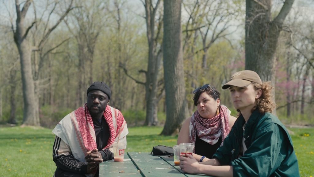 Three university students sit at a picnic table in a park. One man sits on the left - he is Black and wears a red and white keffiyeh. A white woman on the right also wears a keffiyeh. Seated in front of her is a white man in a green jacket and brown baseball cap.