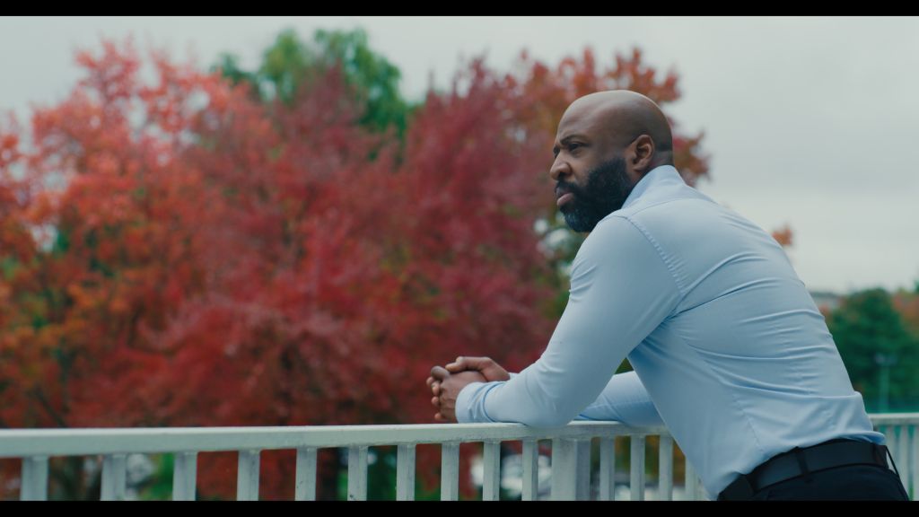 Professor Erec Smith leans on a railing while looking out a university campus. He is a Black man wearing a white collared shirt, and has a shaved head. The leaves on the trees have turned red.
