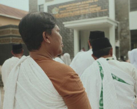 A group of people preparing for Hajj wear white robes. We see the group from behind, with one person centre-frame with shorter hair and a brown sleeved shirt.