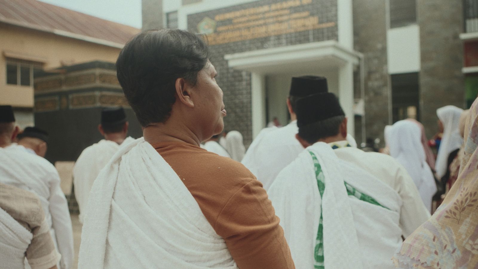 A group of people preparing for Hajj wear white robes. We see the group from behind, with one person centre-frame with shorter hair and a brown sleeved shirt.