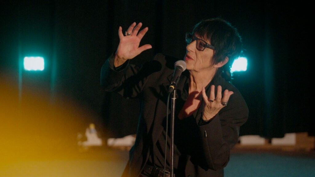 Rocker Carole Pope sings on stage. She is wearing a black collared shirt and black-rimmed glasses. She is a white woman with black hair. There is a blue light in the background.