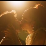 Film subjects Lourenzo and Isis, a transman and transwoman, respectively, share a kiss in a scene with stylish lighting. There is a flood light at the top of the frame.