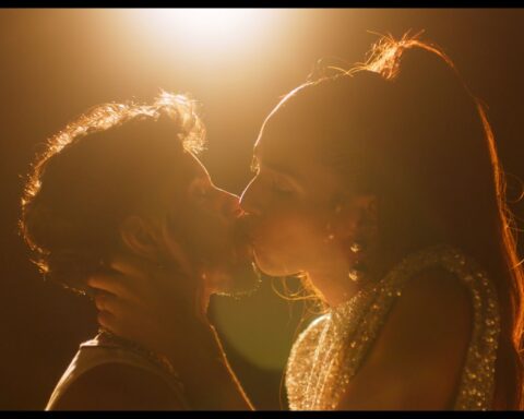 Film subjects Lourenzo and Isis, a transman and transwoman, respectively, share a kiss in a scene with stylish lighting. There is a flood light at the top of the frame.