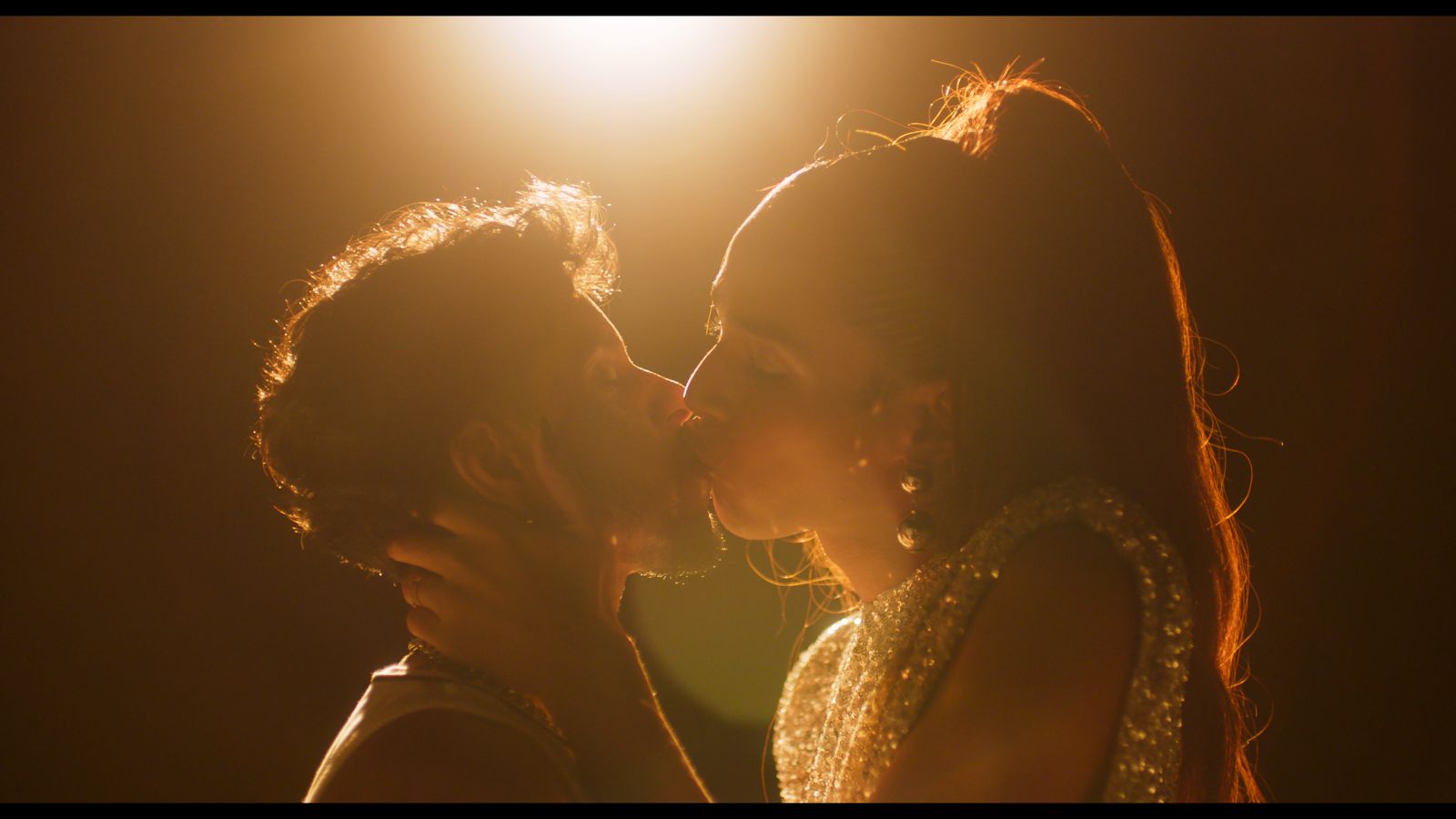 Film subjects Lourenzo and Isis, a transman and transwoman, respectively, share a kiss in a scene with stylish lighting. There is a flood light at the top of the frame.