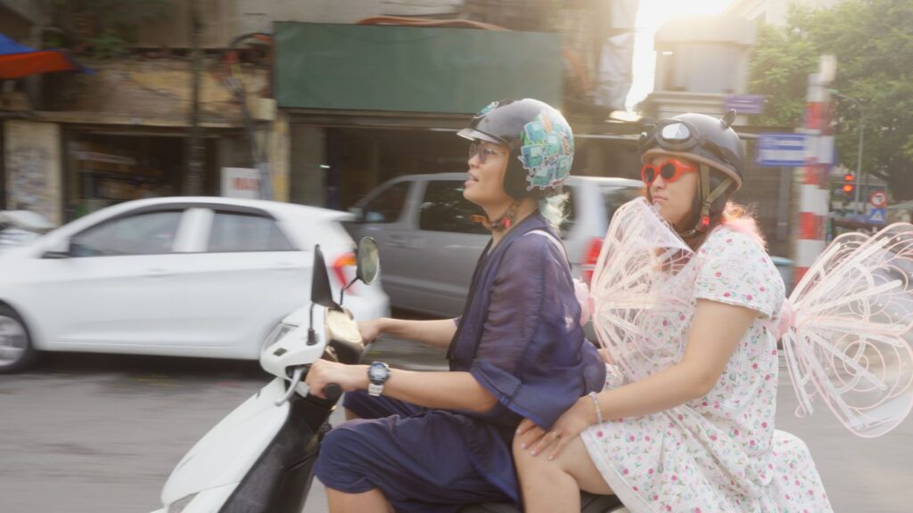 A Vietnamese man rides a scooter while his older sister sits in the back eat. They are dressed fabulously for Pride celebrations.