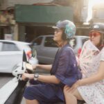 A Vietnamese man rides a scooter while his older sister sits in the back eat. They are dressed fabulously for Pride celebrations.