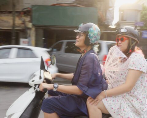 A Vietnamese man rides a scooter while his older sister sits in the back eat. They are dressed fabulously for Pride celebrations.