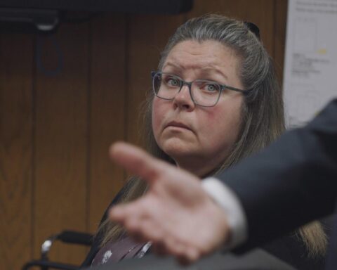 Gail Eastwood-Ritchey, a white woman with glasses and greying light-brown hair, sits in a courtroom and looks in the direction of a man making a speech. The man is partly visible in the foreground of the photo. He is standing up and only his arm and hand are in the shot as he makes a gesture.