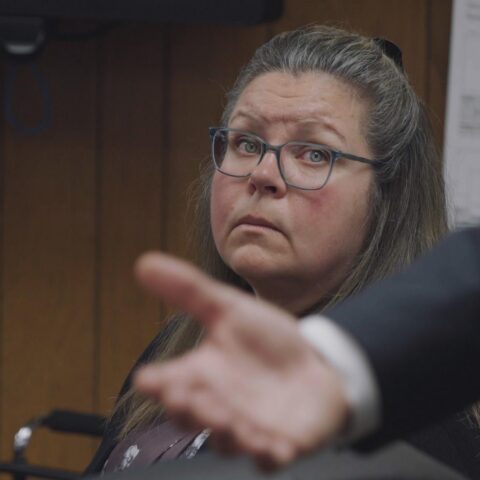 Gail Eastwood-Ritchey, a white woman with glasses and greying light-brown hair, sits in a courtroom and looks in the direction of a man making a speech. The man is partly visible in the foreground of the photo. He is standing up and only his arm and hand are in the shot as he makes a gesture.