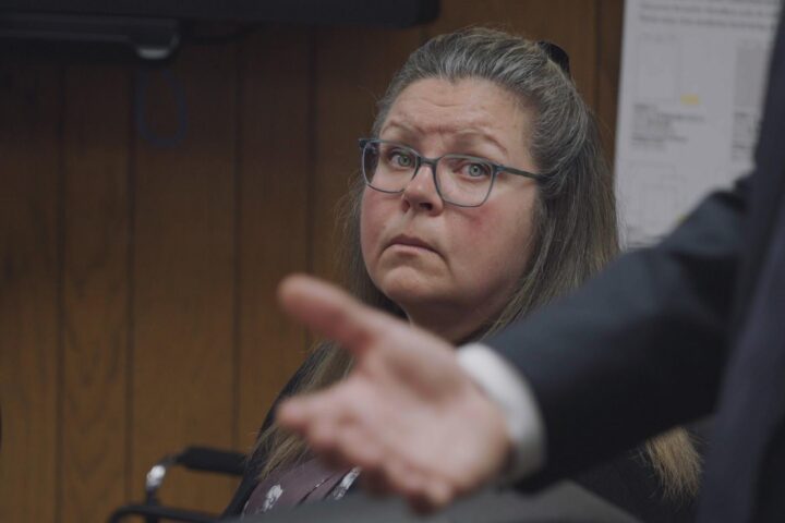 Gail Eastwood-Ritchey, a white woman with glasses and greying light-brown hair, sits in a courtroom and looks in the direction of a man making a speech. The man is partly visible in the foreground of the photo. He is standing up and only his arm and hand are in the shot as he makes a gesture.