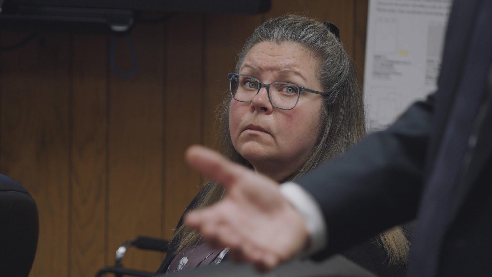 Gail Eastwood-Ritchey, a white woman with glasses and greying light-brown hair, sits in a courtroom and looks in the direction of a man making a speech. The man is partly visible in the foreground of the photo. He is standing up and only his arm and hand are in the shot as he makes a gesture.