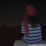 A woman and an animated fox sit with their back to the camera, looking up at a pink crescent moon in the night sky.