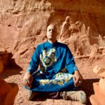 Starseed Arcturus Ra meditates in a desert. He is a bald Brown man and is wearing a blue robe with yellow markings. The terrain surrounding him is red sand and rock.