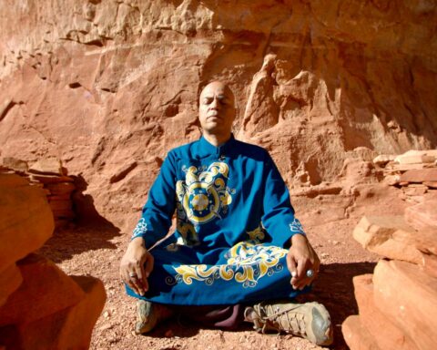Starseed Arcturus Ra meditates in a desert. He is a bald Brown man and is wearing a blue robe with yellow markings. The terrain surrounding him is red sand and rock.