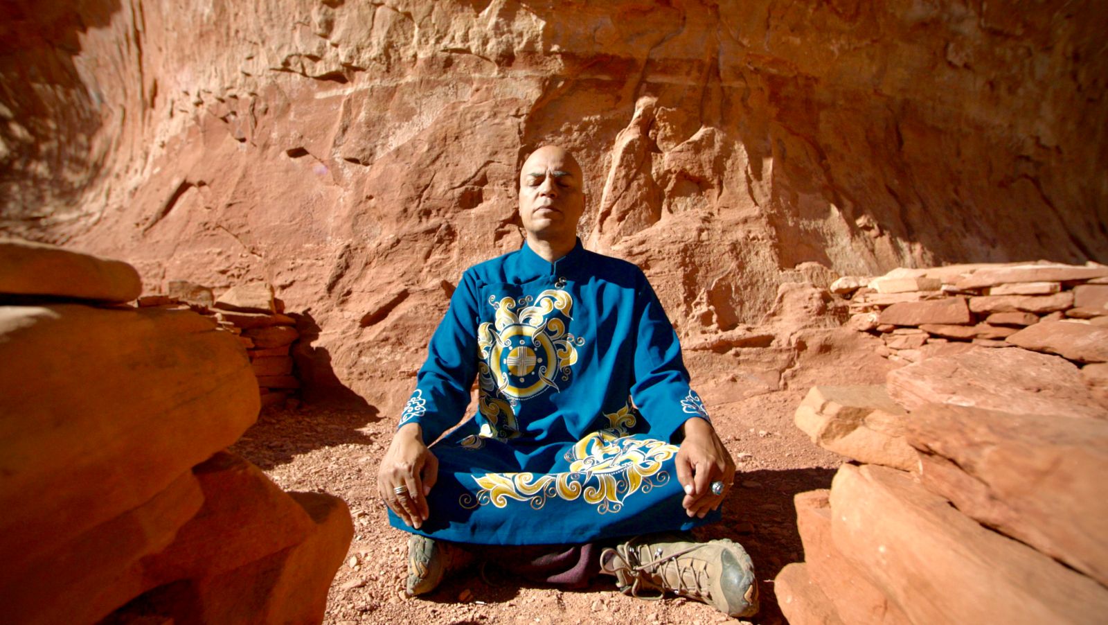Starseed Arcturus Ra meditates in a desert. He is a bald Brown man and is wearing a blue robe with yellow markings. The terrain surrounding him is red sand and rock.