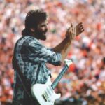 An archival photo of Kenny Loggins in concert. He is clapping and has a guitar strapped over his shoulder. He is a white man with brown hair and is wearing a blue and white plaid shirt. A crowded audiences fills the background.