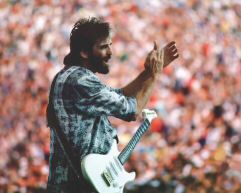 An archival photo of Kenny Loggins in concert. He is clapping and has a guitar strapped over his shoulder. He is a white man with brown hair and is wearing a blue and white plaid shirt. A crowded audiences fills the background.