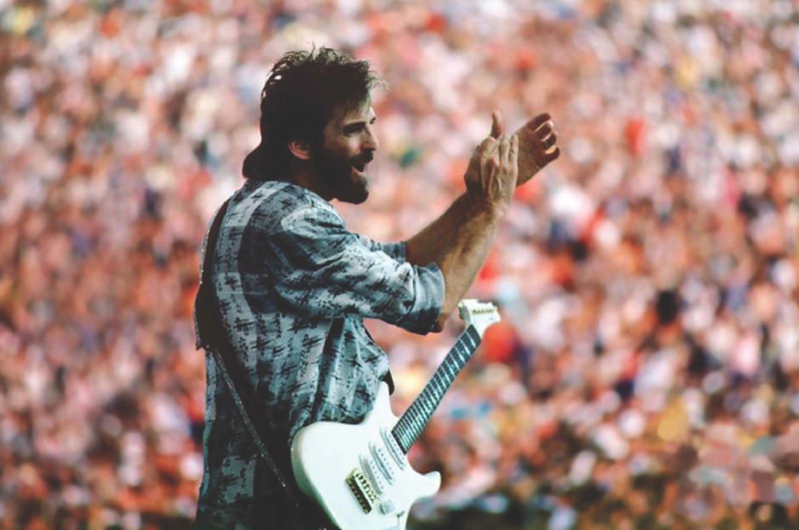 An archival photo of Kenny Loggins in concert. He is clapping and has a guitar strapped over his shoulder. He is a white man with brown hair and is wearing a blue and white plaid shirt. A crowded audiences fills the background.