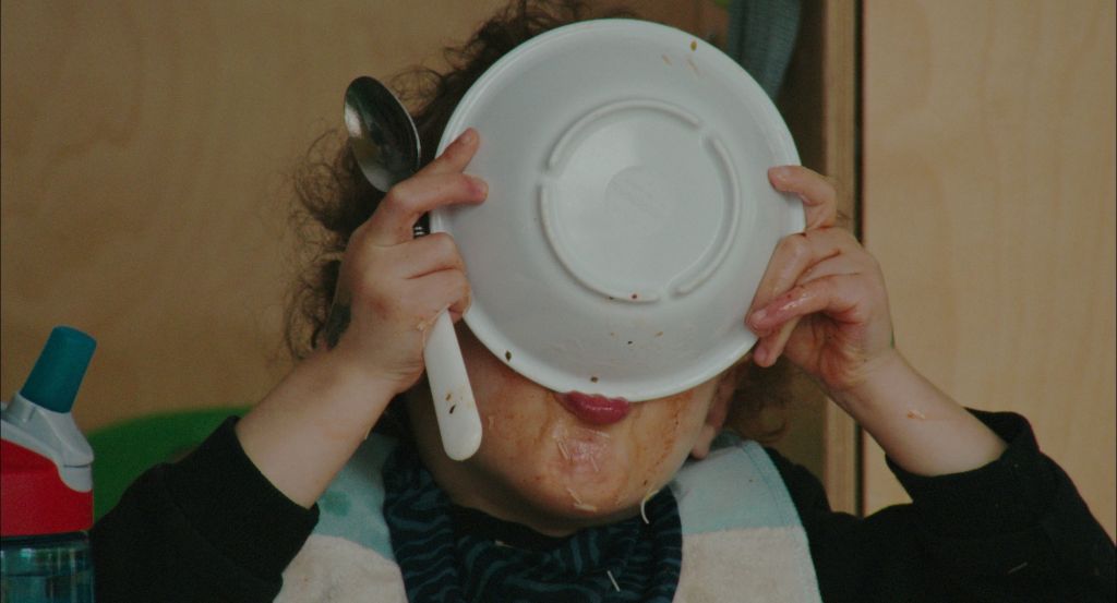 A kid holds a bowl of soup overtop its face. They are making a mess.