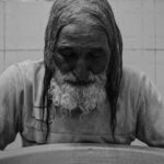 A black and white photo of an elderly man with a busy beard. He stands before a very large bowl and has his arms outstretched around it.