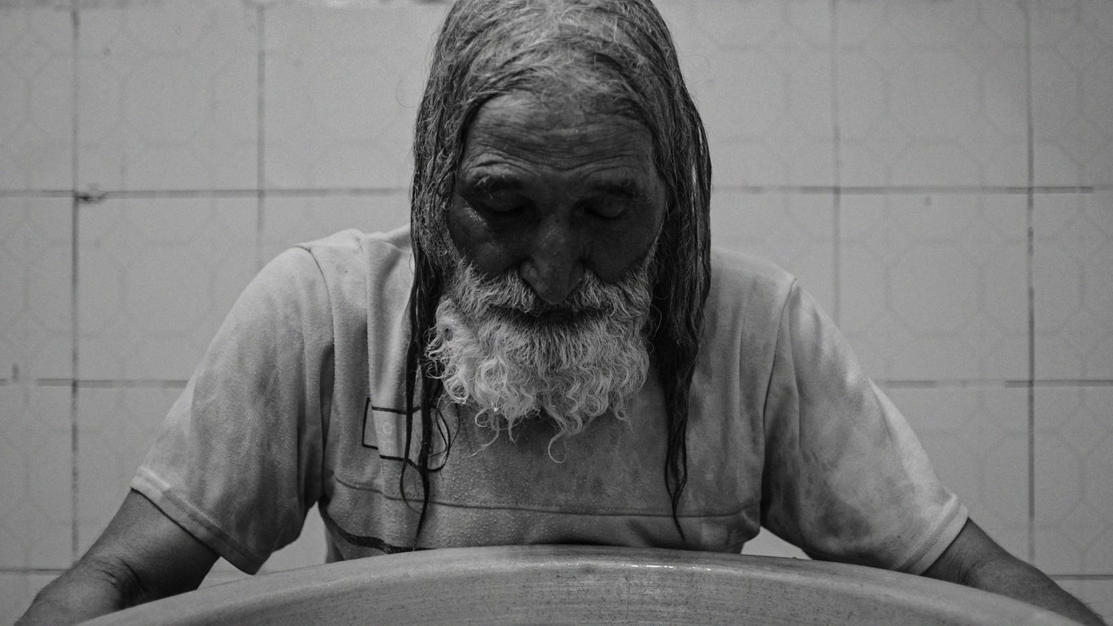 A black and white photo of an elderly man with a busy beard. He stands before a very large bowl and has his arms outstretched around it.