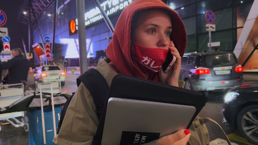 Journalist Olga Churakova stands on a street in Moscow and speaks on her phone. She is holding her laptop and wearing a face mask. She has an apprehensive expression.