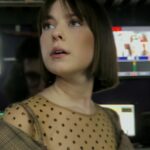 A Russian woman in her 20s stands in a broadcasting studio. She looks up at monitors with an anxious expression. There are several TV screens behind her.