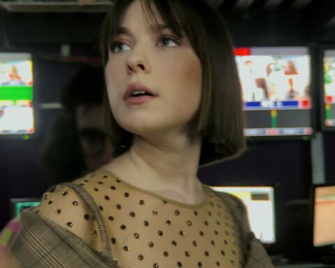 A Russian woman in her 20s stands in a broadcasting studio. She looks up at monitors with an anxious expression. There are several TV screens behind her.