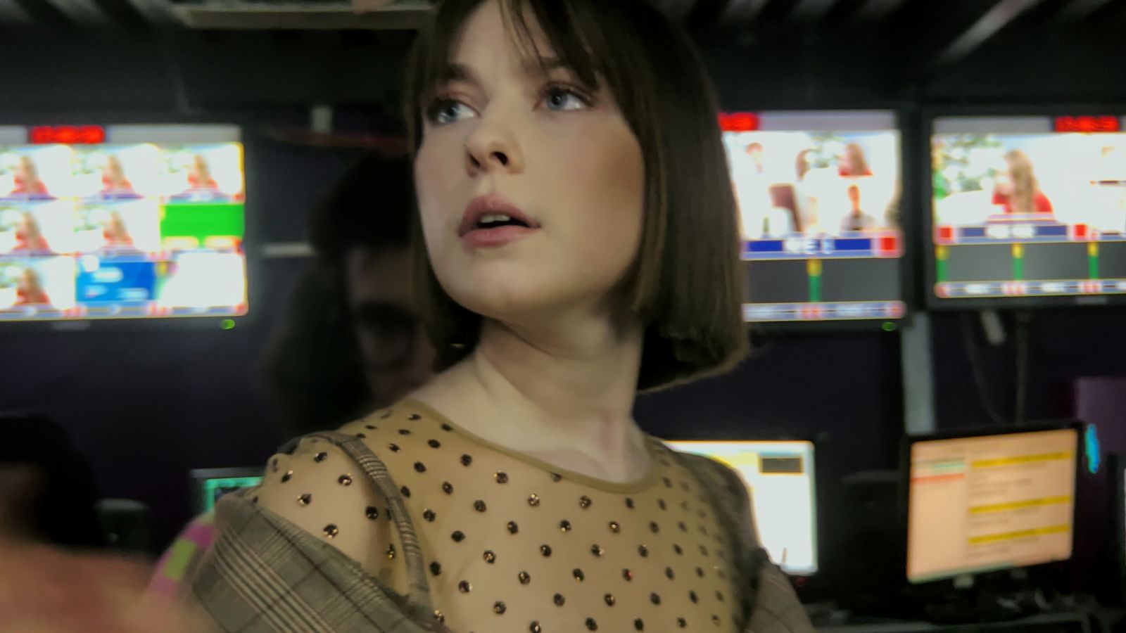 A Russian woman in her 20s stands in a broadcasting studio. She looks up at monitors with an anxious expression. There are several TV screens behind her.
