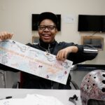Nekai Foster holds a banner that reads "Congratulations on your discharge, Nekai." He is a young Black man seated a a table. He wears a black track suit, ballcap, and glasses. He is smiling.