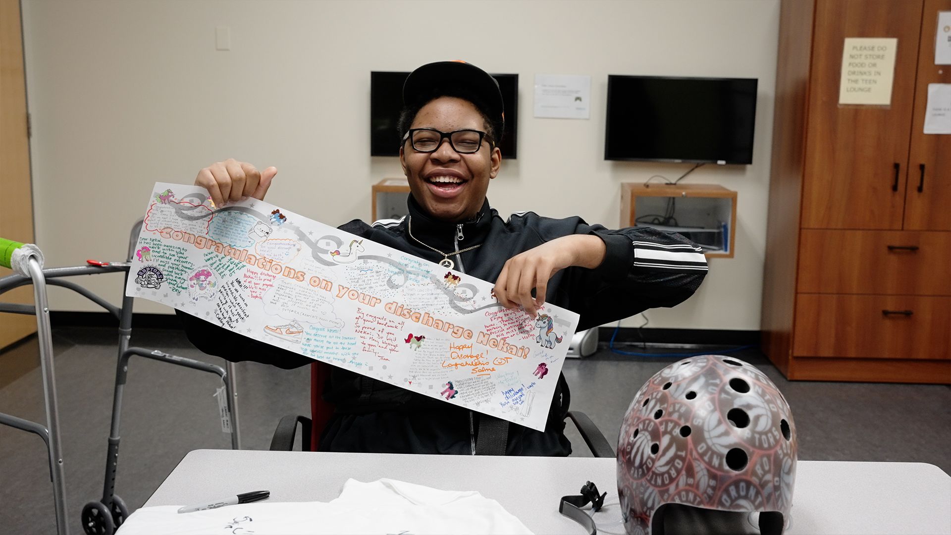 Nekai Foster holds a banner that reads "Congratulations on your discharge, Nekai." He is a young Black man seated a a table. He wears a black track suit, ballcap, and glasses. He is smiling.