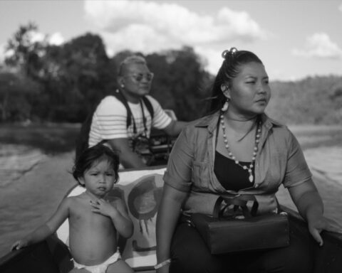 A black and white image of three Wayana on a boat in the Lawa River, South America. The travellers include an adult mother, a small child in a diaper, and an older man at the back of the boat steering the motor.