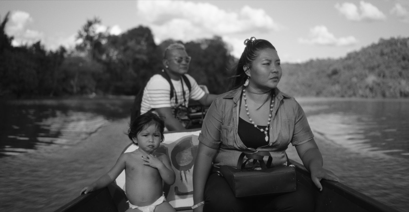 A black and white image of three Wayana on a boat in the Lawa River, South America. The travellers include an adult mother, a small child in a diaper, and an older man at the back of the boat steering the motor.