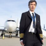 Actor Nathan Fielder stands on an airport runway. He is a white man with brown hair. He is wearing a pilot's uniform with a suit and tie. An airplane is in the background.