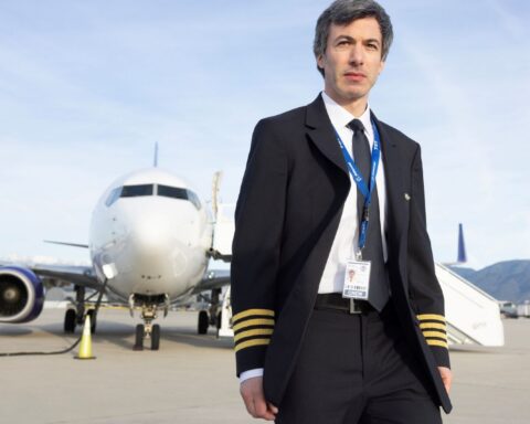 Actor Nathan Fielder stands on an airport runway. He is a white man with brown hair. He is wearing a pilot's uniform with a suit and tie. An airplane is in the background.