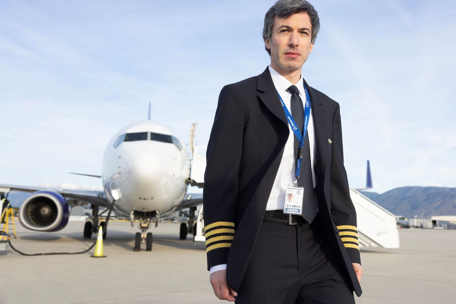 Actor Nathan Fielder stands on an airport runway. He is a white man with brown hair. He is wearing a pilot's uniform with a suit and tie. An airplane is in the background.