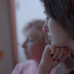 A young woman is pictured in close-up. She is white and has brown hair. Her head rests on her hand beneath her chin and she has a thoughtful look.