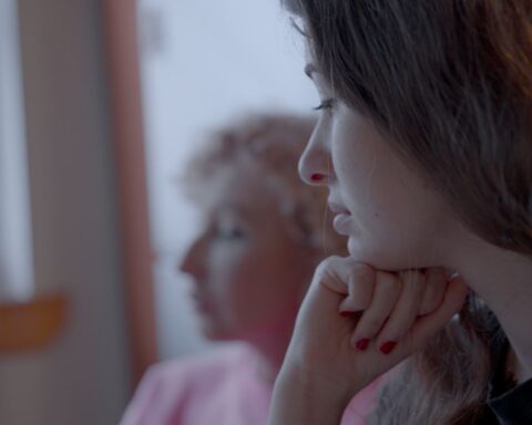 A young woman is pictured in close-up. She is white and has brown hair. Her head rests on her hand beneath her chin and she has a thoughtful look.