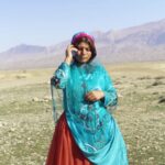 An Iranian woman stands in a field talking on a cell phone. She is wearing a red and blue robe with a red veil.