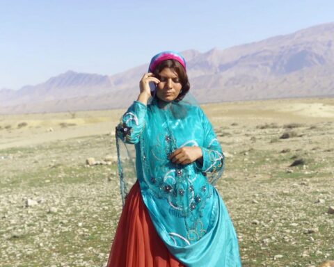 An Iranian woman stands in a field talking on a cell phone. She is wearing a red and blue robe with a red veil.