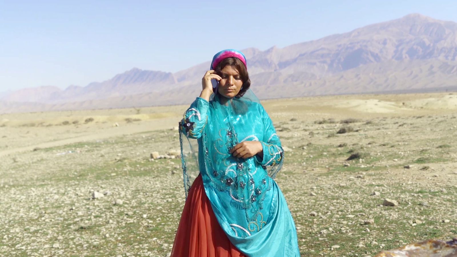 An Iranian woman stands in a field talking on a cell phone. She is wearing a red and blue robe with a red veil.