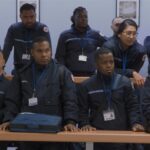 A group of adult students in a prison guard classroom stand attentively while listening to a speaker outside the frame. The racially diverse group of students are all wearing the same navy blue uniform with a white badge on a lanyard around their necks.