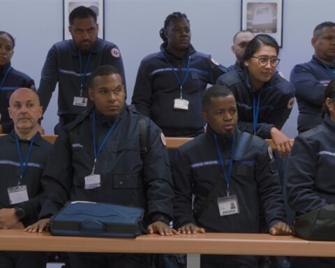 A group of adult students in a prison guard classroom stand attentively while listening to a speaker outside the frame. The racially diverse group of students are all wearing the same navy blue uniform with a white badge on a lanyard around their necks.